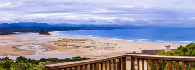 Elevated panorama of Lookout Beach and the Keurbooms Estuary in Plettenberg Bay, showing tidal channels, sandbanks, ocean surf and distant mountain ranges.