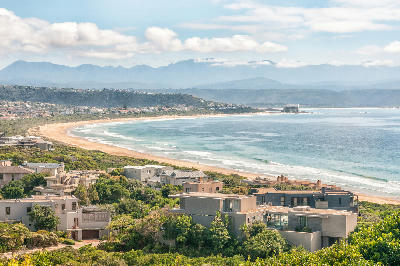 Lookout Beach in Plettenberg Bay with a sweeping sandy shoreline, coastal homes, Beacon Isle in the distance and mountains beyond the bay.