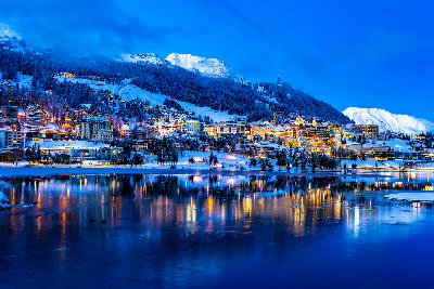 St. Moritz town at night with illuminated buildings reflected in Lake St. Moritz, surrounded by snow-covered slopes and mountains in the Upper Engadin, Switzerland.