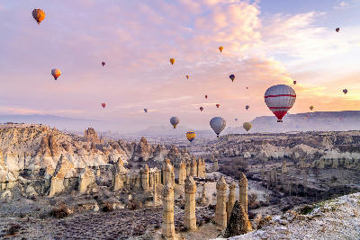Cappadocia with multiple hot air balloons floating above volcanic rock formations during early daylight conditions.