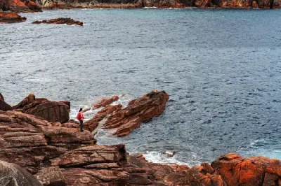 Person standing among layered red granite rocks beside the sea at Sleepy Bay, Freycinet National Park, Tasmania.