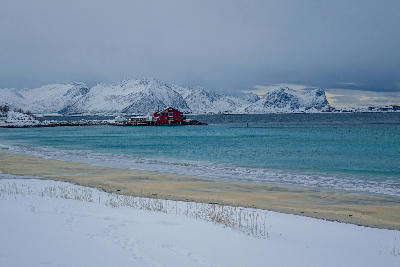 Snow-covered Bøvær beach on Senja with turquoise water, red coastal building, and mountains in winter, northern Norway