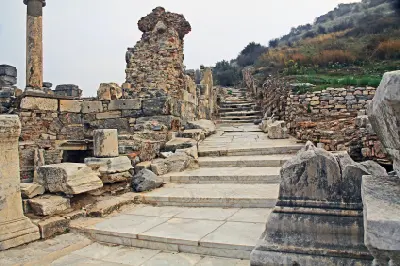 Marble steps climb a narrow side street off Curetes Street, surrounded by weathered columns, stone walls, and ruins in ancient Ephesus.