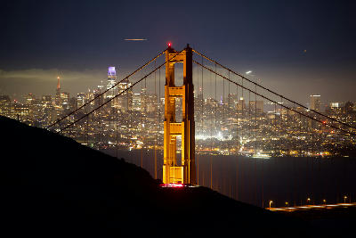Golden Gate Bridge tower illuminated at night with San Francisco skyline lights in the background