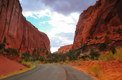 Burr Trail Road curves between towering red sandstone cliffs and desert vegetation beneath dramatic clouds in Long Canyon.