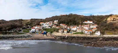 Aerial view from the sea of Runswick Bay, showing red-roofed cottages on the hillside, a small sandy beach, stone harbour walls and waves along the North Yorkshire coast.
