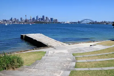 View from the Bradleys Head foreshore across Sydney Harbour towards the CBD skyline, Opera House and Harbour Bridge, with a stone jetty in the foreground.