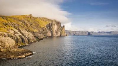 Trøllkonufingur sea stack, known as the Witch’s Finger, standing off the rugged cliffs of Vágar Island with Atlantic waters below and low cloud along the coastline in the Faroe Islands.