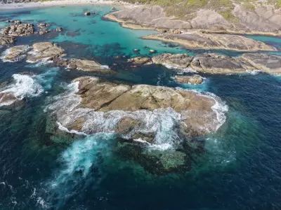 High-angle aerial perspective of waves creating white sea foam as they sweep over large boulders in a clear turquoise sea.