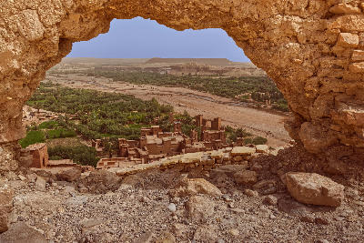 View through a ruined wall above Aït Benhaddou showing the earthen kasbah village, oasis landscape, and river valley in Morocco.