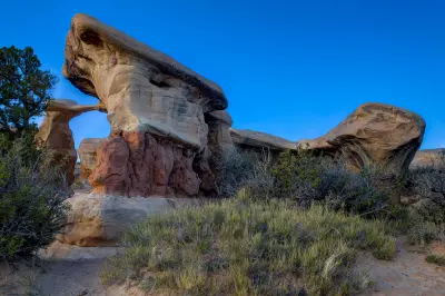 Metate Arch rises among large sculpted sandstone formations and desert vegetation beneath a deep blue sky in Devil’s Garden.