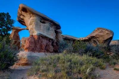 Metate Arch rises among large sculpted sandstone formations and desert vegetation beneath a deep blue sky in Devil’s Garden.