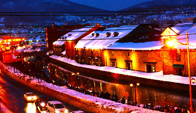 Snow-covered warehouses illuminated at night along Otaru Canal with warm lights reflecting in the water, Hokkaido, Japan.