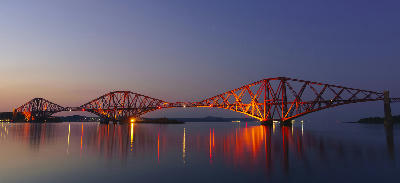 The Forth Bridge illuminated at dusk with reflections in the Firth of Forth near South Queensferry in Central Scotland.