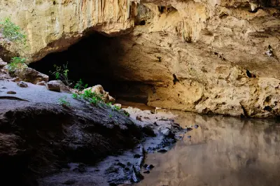 Shallow creek water flows through the dark limestone passage of Tunnel Creek, surrounded by weathered cave walls and sandy banks.