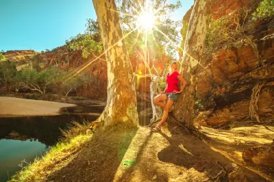 A woman in a red shirt stands next to massive white-barked eucalyptus trees, highlighting the grand scale of the towering red cliffs and sun-drenched outback nature landscape surrounding the waterhole.