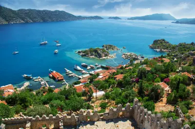 Panoramic view from Simena Castle in Kaleköy, Kekova, Turkey, overlooking red-roofed village houses, a small harbour, rocky islands, sailing boats and vivid turquoise Mediterranean water.