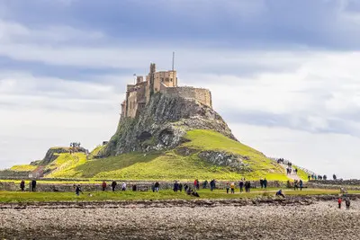 Lindisfarne Castle overlooking a grassy rocky hill on Holy Island, with visitors walking beside stone walls and a rocky shoreline in the foreground.