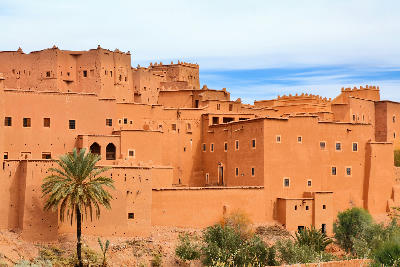 Adobe walls and fortified towers of Kasbah Taourirt in Ouarzazate, Morocco, surrounded by palms and desert vegetation.