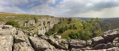 Panoramic view from the limestone pavement above Malham Cove, showing layered limestone cliffs, walkers and rolling green countryside in the Yorkshire Dales.