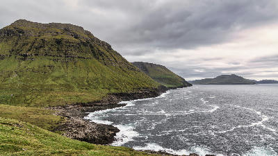 Rugged coastal cliffs and green mountains along Norðradalur on Streymoy, Faroe Islands under dramatic cloudy skies.