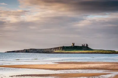 Dunstanburgh Castle on a rocky headland overlooking Embleton Bay, with exposed sandbanks, shallow tidal water and a softly illuminated cloudy sky.