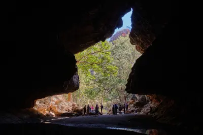 Visitors silhouetted inside the vast limestone cavern at Tunnel Creek, looking toward the sunlit forest beyond the cave entrance.