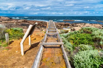 Historic Cape Leeuwin Water Wheel among granite rocks and coastal vegetation beside the ocean near Cape Leeuwin, Western Australia.