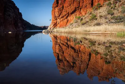 Red-orange rock formations reflect in the calm, glassy water of the Glen Helen Gorge waterhole beneath a clear blue sky.