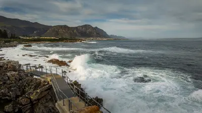 Scenic viewing platform along the Hermanus Cliff Path with crashing ocean waves, rugged coastal rocks, Walker Bay, and mountain scenery in the Overberg region of South Africa.