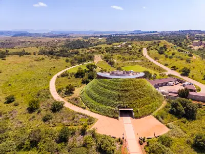 Aerial view of the Maropeng Visitor Centre, a grass-covered mound-shaped building surrounded by walking paths, open grasslands and rolling hills in the Cradle of Humankind, Gauteng, South Africa.