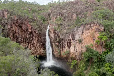 Powerful white waterfall dropping between towering red-brown sandstone cliffs into a dark plunge pool surrounded by green woodland at Tolmer Falls, Litchfield National Park, Northern Territory, Australia.