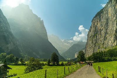 Path through green meadows on the Lauterbrunnen Valley floor with steep cliff walls and distant alpine peaks under morning light, Switzerland.