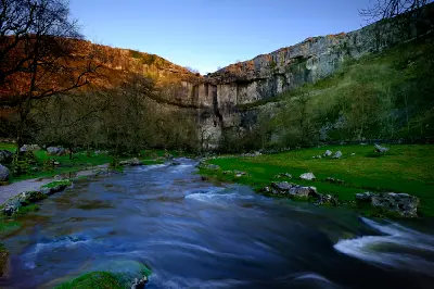 Flowing stream beneath the curved limestone cliffs of Malham Cove, with bare winter trees and warm sunlight on the upper rock face in the Yorkshire Dales.