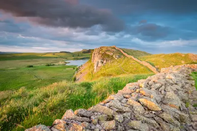 Hadrian's Wall winding over a rocky ridge above Crag Lough and Highshield Crags, with green moorland and dramatic evening clouds in Northumberland, England.