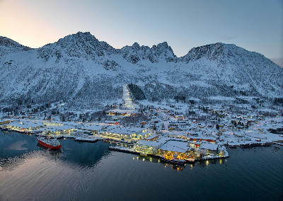 Aerial view of Senjahopen harbour Senja Norway showing fishing boats, industrial buildings and village below snow covered mountain slopes at dusk.