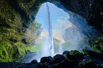 Kvernufoss waterfall on Iceland’s South Coast, seen from inside a cave-like opening with green moss-covered cliffs and water falling into a rocky basin below.