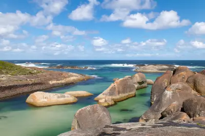 Rounded grey-brown boulder rocks emerge from clear, shallow green ocean water bordered by low coastal hills.