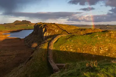 Hadrian's Wall curving along the rocky escarpment above Crag Lough in Northumberland, England, with golden evening light and a faint rainbow.