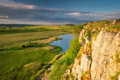Golden sunlight on the rocky cliffs of Highshield Crags overlooking Crag Lough, with green moorland and dramatic clouds in Northumberland, England.