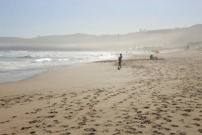 An angler stands on Wilderness Beach beside the Indian Ocean, with surf rolling onto the sand beneath hazy, wind-blown coastal conditions.