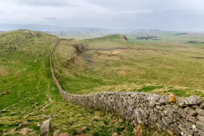 Ancient stonework of Hadrian's Wall curving over grassy hills in Northumberland beneath a pale, overcast sky.