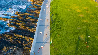Aerial view of people walking along the Sea Point Promenade beside the rocky Atlantic coastline and park lawns in Cape Town, South Africa.