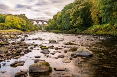 River-level view towards the stone arches of Lambley Viaduct, with shallow flowing water, mossy rocks and autumn trees along the River South Tyne in Northumberland.