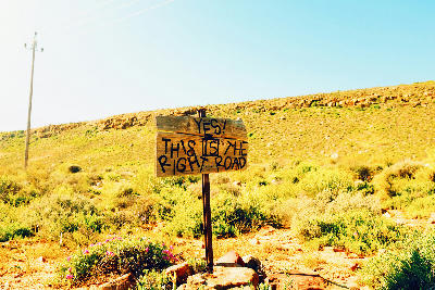 Roadside sign in the remote Namaqualand landscape of the Northern Cape, South Africa, surrounded by semi-arid vegetation and open countryside.