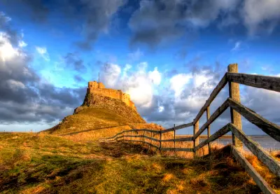 Lindisfarne Castle standing on a steep rocky hill on Holy Island, Northumberland, with a wooden fence leading towards the castle beneath dramatic blue and grey winter clouds.