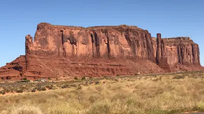 Large sandstone mesa in Monument Valley Arizona with sheer cliffs rising above desert grassland and small vehicles at base showing scale