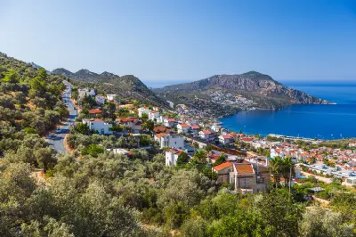 Kalkan’s whitewashed houses and red-tiled roofs spread across green hillsides above a brilliant blue Mediterranean bay, framed by a rugged rocky headland.