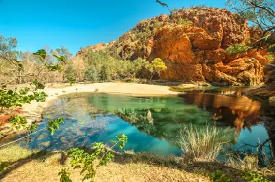 A tranquil, clear green waterhole reflects towering red-orange cliffs under a bright blue sky, with a sandy bank and eucalyptus trees surrounding the water.