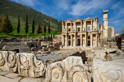 Library of Celsus rising among the ruins of Ephesus, with carved marble blocks in the foreground and a green hillside behind.